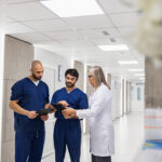 Three medical professionals standing in a bright hospital hallway reviewing patient information on digital tablets, collaborating on clinical workflow.