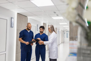Three medical professionals standing in a bright hospital hallway reviewing patient information on digital tablets, collaborating on clinical workflow.